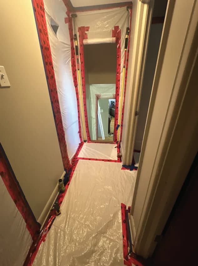 Polyethylene containment sealed across a residential hallway and doorway, taped at every seam and held by adjustable support poles, with floor protection running through to the room beyond