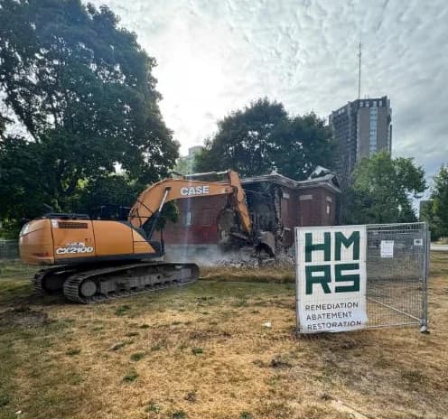 Excavator working on a fire-damaged brick building behind temporary site fencing carrying an HMRS site sign