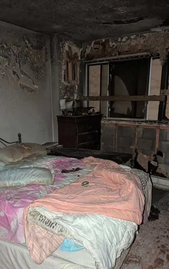 Bedroom after a fire, with the ceiling burned through, soot and blistered paint across the walls, and the window frame charred out to the block behind it